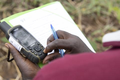 View over the shoulder, right hand holds a pen over a clipboard, left had holds a GPS tracking device.